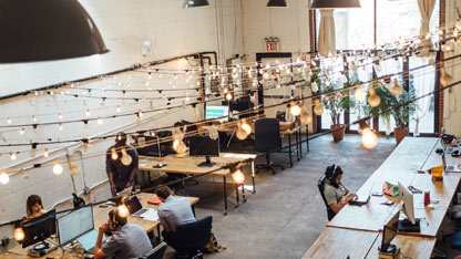 Biuro w układzie otwartej przestrzeni z pracującymi ludźmi przy biurkach, światełkami na linkach i naturalnym światłem z okien.